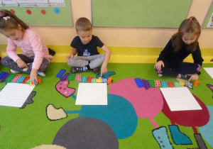 Troje dzieci siedzi na dywanie przed płytką i z ułożonymi klockami w szeregu według wzrastającej wartości. Dzieci przeliczają oczka na klockach.
